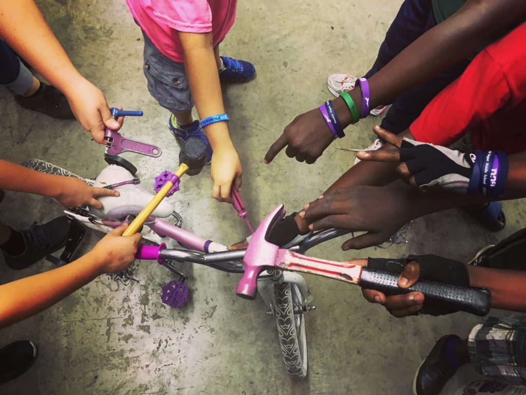Nine children's arms and hands extend toward the middle, holding various tools over a kids' bicycle